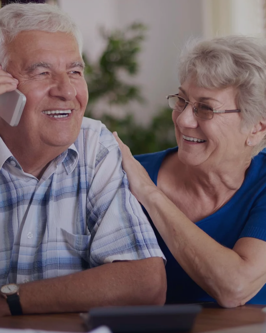 A smiling senior couple at home, one of them on the phone with Illness Support Group